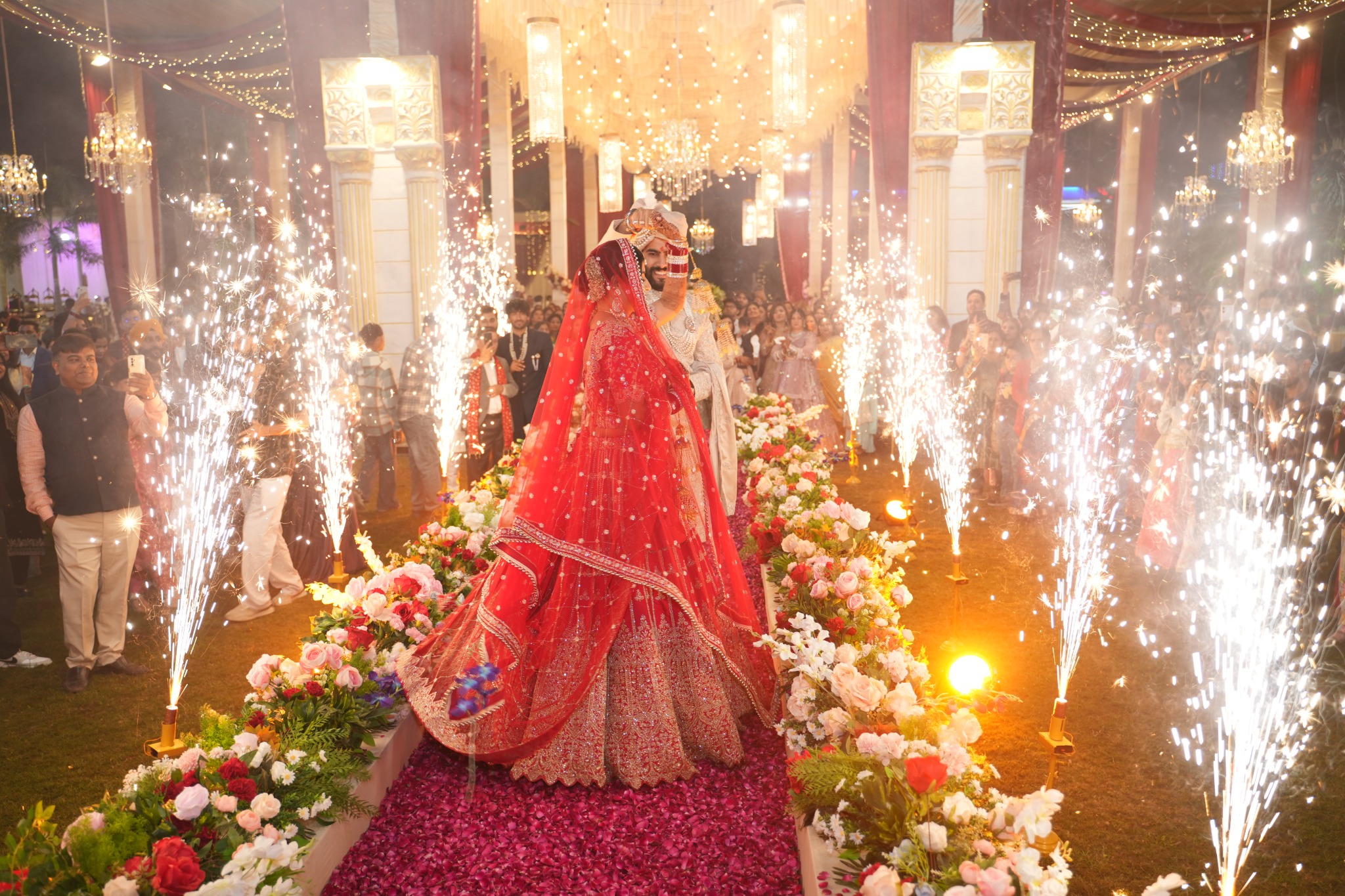 A bride and groom celebrating in a floral wedding aisle framed by sparklers and chandeliers